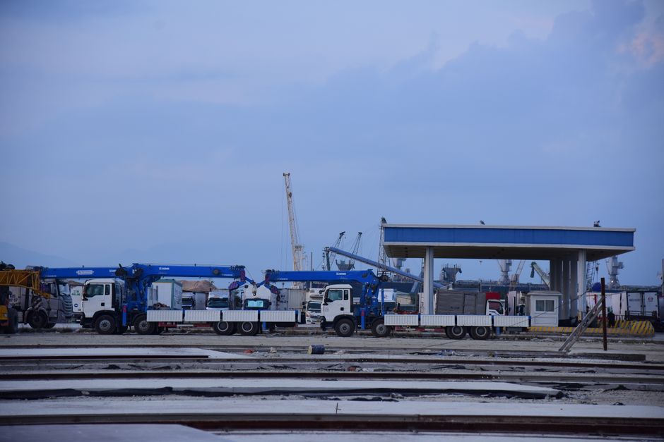 An industrial shipping yard featuring cranes and trucks under a partly cloudy sky.