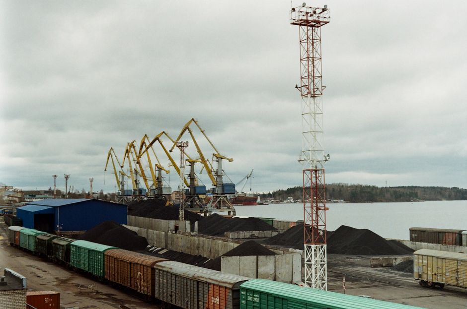A bustling Russian port featuring cargo trains and cranes along the waterfront under a cloudy sky.